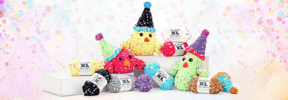A colorful display of plush, crocheted bird toys made from Premier Parfait XL Pom Party yarn. The birds are crafted in bright hues like red, yellow, green, and purple, each wearing a playful, cone-shaped party hat with pom-pom tips. Surrounding the birds are several skeins of the plush, multicolored yarn in shades of black, gray, yellow, purple, blue, and brown. The background features a festive pastel pattern with bubbles and marbled textures.