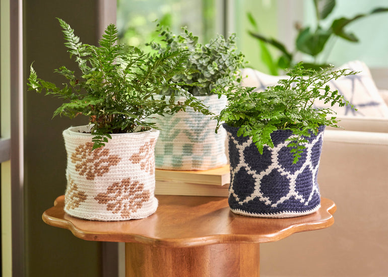 Three crochet plant cozies with geometric and floral patterns in navy & white, tan & white, and mint & white, sitting on wooden flower shaped table with an arm chair in background.